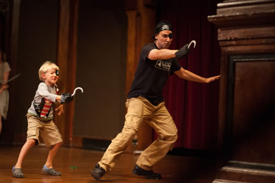 Young boy in eye patch and hooked right hand, wearing t-shirt and cargo shorts, and older man in black skull cap eyepatch, hook on right hand, black t-shirt and cargo pants stand poised to attack